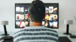 Mężczyzna w bluzie w paski ogląda telewizor. Zastanawia się, jak uniknąć kary za niepłacenie abonamentu RTV, szukając alternatywnych rozwiązań.