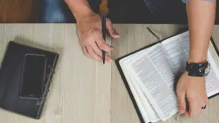 Otwarta Biblia, notatnik i telefon na drewnianym stole. Dłoń z długopisem przygotowuje się do codziennego rachunku sumienia.