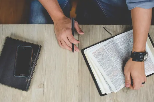 Otwarta Biblia, notatnik i telefon na drewnianym stole. Dłoń z długopisem przygotowuje się do codziennego rachunku sumienia.