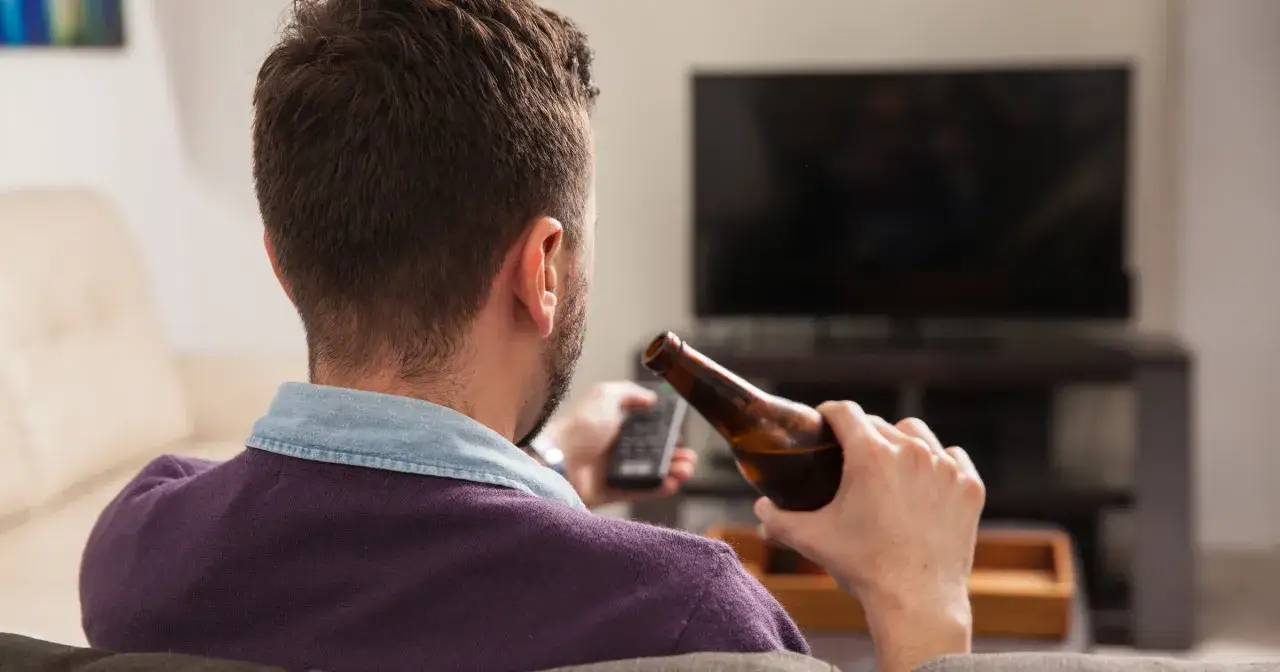 Mężczyzna siedzi na kanapie, trzymając pilota i butelkę piwa. Codzienne picie piwa to jego sposób na relaks przed telewizorem.