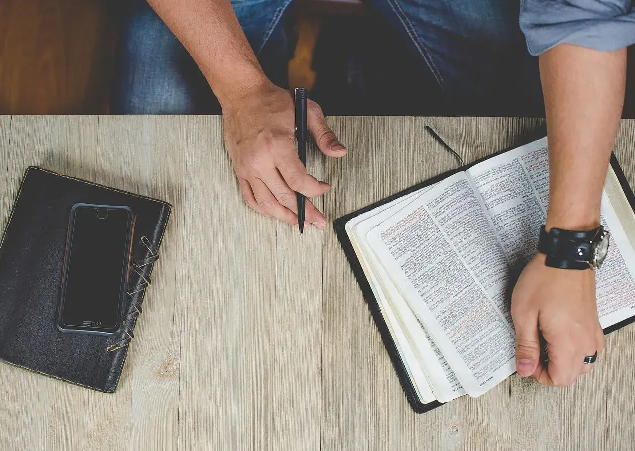 Otwarta Biblia, notatnik i telefon na drewnianym stole. Dłoń z długopisem przygotowuje się do codziennego rachunku sumienia.