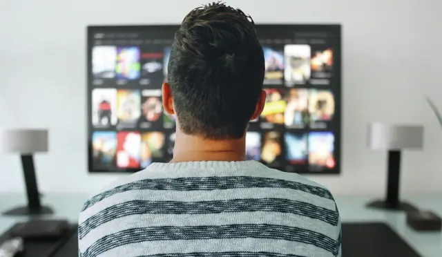 Mężczyzna w bluzie w paski ogląda telewizor. Zastanawia się, jak uniknąć kary za niepłacenie abonamentu RTV, szukając alternatywnych rozwiązań.